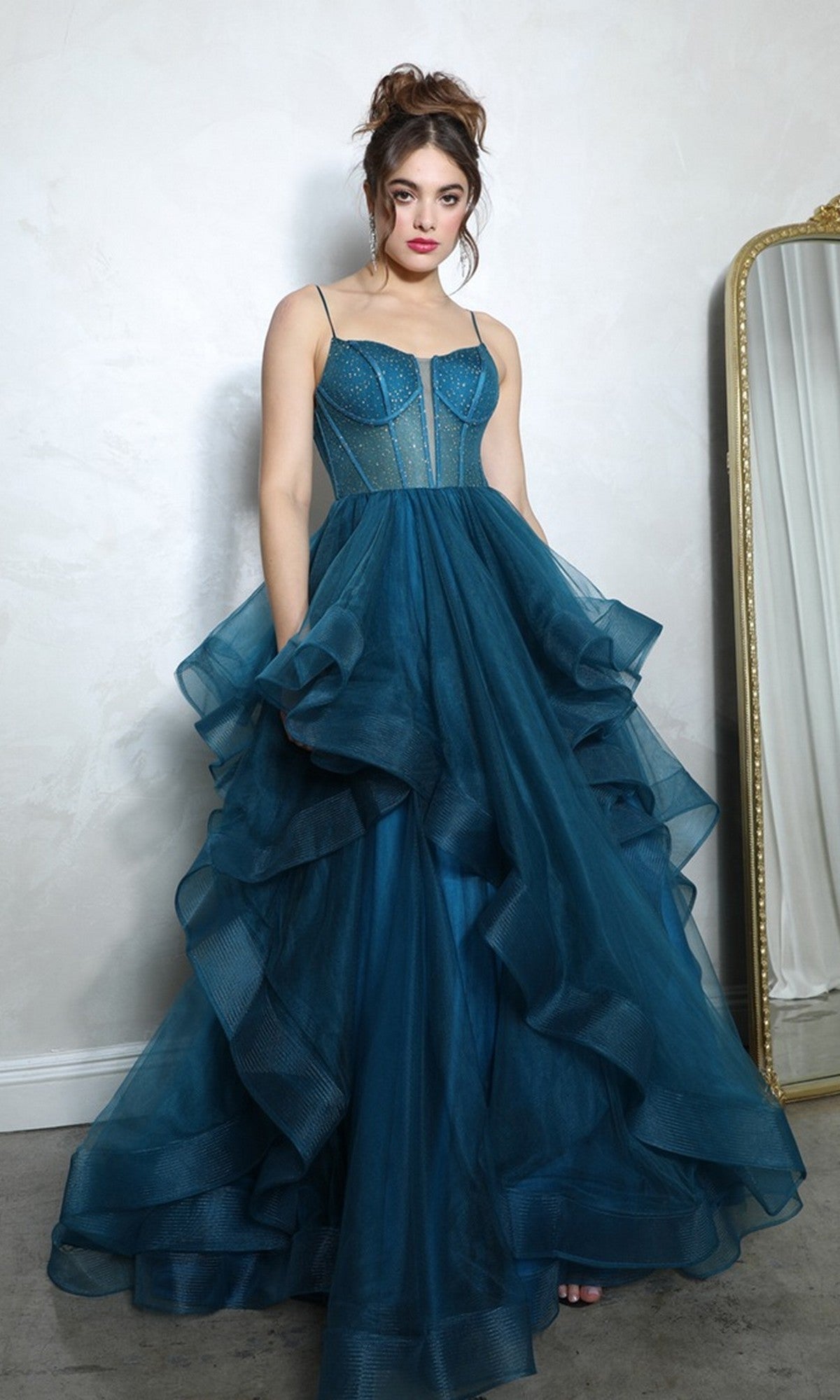 A woman stands indoors in the Eureka 9950 Long Prom Dress, featuring a teal beaded corset bodice and spaghetti straps, next to a large ornate mirror against a light-colored wall.